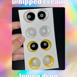 Op de afbeelding: Een vel met zes knutselogen in verschillende ontwerpen. De bovenste twee paren zijn zwart met glitter, het middelste paar is wit met glitter en de onderste twee paren zijn geel met glitter. De tekst "whipped cream" en "lemon drop" staan bovenaan en onderaan.