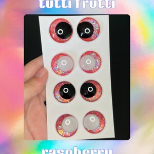 Op de afbeelding: Een vel frambooskleurige knutselogen met zwarte pupillen en glitteraccenten. De tekst "tutti frutti" staat bovenaan en "raspberry" onderaan. De Instagram-handle @raylouisecrochet is ook zichtbaar.