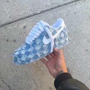 May include: A light blue denim sneaker with a white swoosh logo and white laces. The sneaker has a checkered pattern and frayed edges.