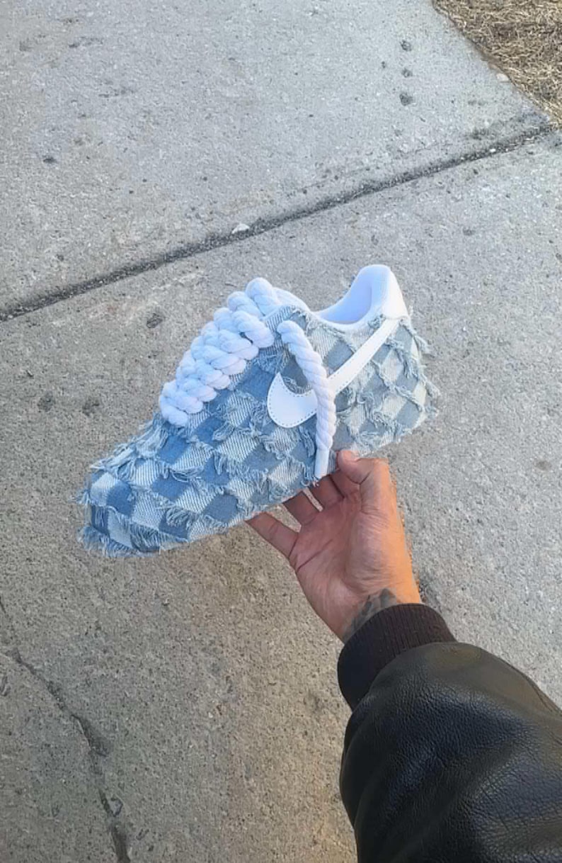 May include: A light blue denim sneaker with a white swoosh logo and white laces. The sneaker has a checkered pattern and frayed edges.