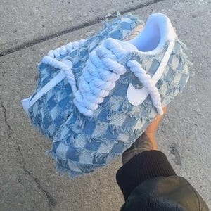 May include: Blue and white checkered denim sneakers with white laces and a white swoosh logo. The laces are tied in a unique way, with the ends of the laces forming a rope-like design.