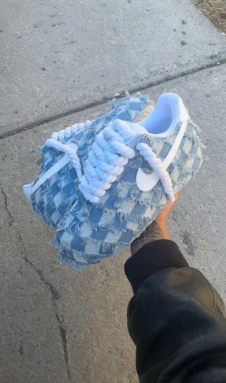 May include: Blue and white checkered denim sneakers with white laces and a white swoosh logo. The laces are tied in a unique way, with the ends of the laces forming a rope-like design.