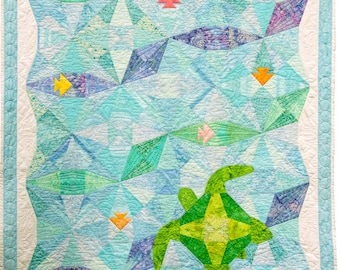 May include: A quilt with a blue background and a green sea turtle in the center. The quilt is made up of geometric shapes in shades of blue, green, and purple. There are also small yellow and red shapes scattered throughout the quilt.