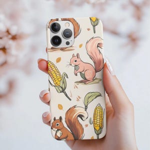 Puede incluir: Funda de teléfono con un diseño otoñal. El diseño presenta un patrón repetido de ardillas, maíz y hojas en tonos marrones, rosas y amarillos sobre un fondo crema. La funda está sujeta por una mano.