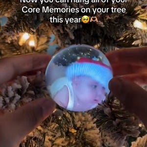 May include: A clear, spherical Christmas ornament displaying a photo of a person wearing a Santa hat. The ornament is held in front of a snow-covered Christmas tree. Text on the image reads, "Now you can hang all of your Core Memories on your tree this year."