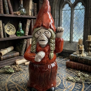 Peut inclure: Une figurine en céramique représentant une créature au visage squelettique, aux longs cheveux tressés et portant un chapeau rouge pointu. Le personnage porte un haut rouge à manches longues et une jupe assortie, un bras levé en geste.