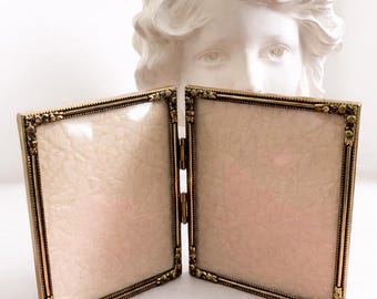 May include: A hinged, double-sided photo frame with a gold-colored, ornate border. The frame's interior displays a pale pink, textured surface. A white bust of a classical figure is in the background.
