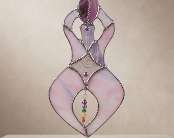 May include: A stained glass suncatcher shaped like a stylized figure in shades of pale pink and purple. The piece features a dark metal frame, a central spiral wire detail, and a vertical strand of small, multi-colored beads hanging in the center.