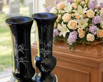 May include: Two black decorative vases with etched silver designs. The vases are cylindrical with a flared top and a bulbous base. A floral arrangement of roses, lilies, and other flowers sits in the background. The vases are on a wooden surface.