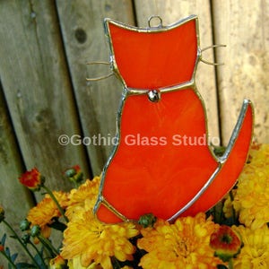 May include: Orange stained glass cat suncatcher with a silver outline. The cat is sitting with its tail curled around its body. The suncatcher is hanging in front of a wooden fence and yellow flowers.