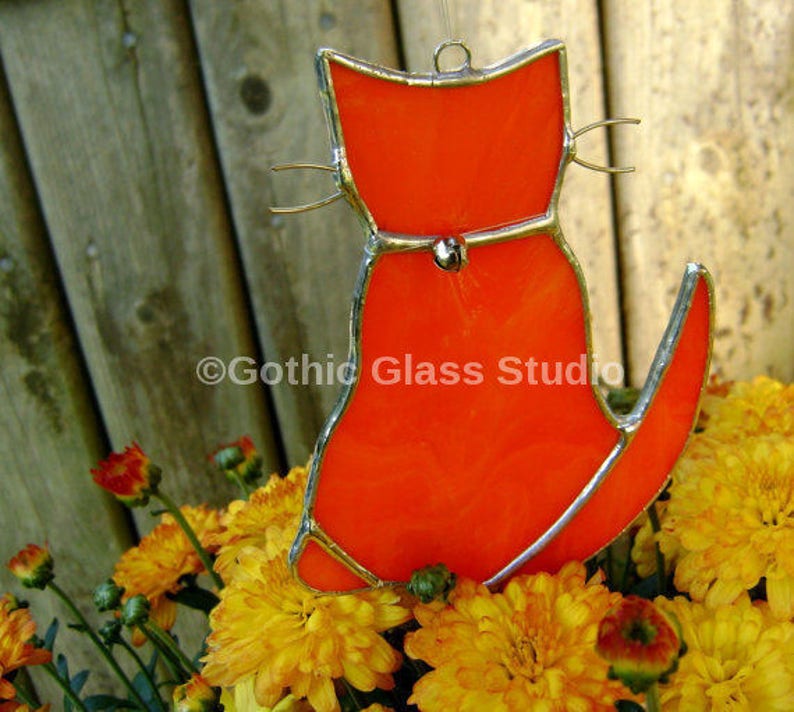 May include: Orange stained glass cat suncatcher with a silver outline. The cat is sitting with its tail curled around its body. The suncatcher is hanging in front of a wooden fence and yellow flowers.