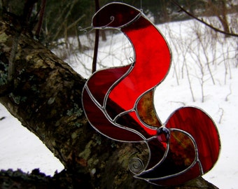 May include: A stained glass snake ornament crafted from translucent red glass panels held together by silver-toned metal solder lines. The snake features a curved, S-shaped body, a small coiled wire tongue, and a dark circular glass accent at the tail.