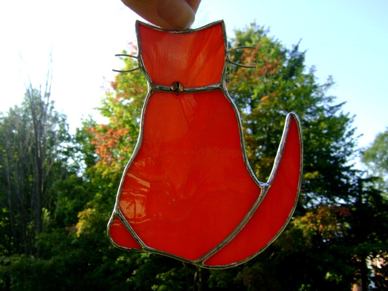 May include: A stained glass cat figurine with a bright orange body and silver accents. The cat is facing to the right and has a long tail.