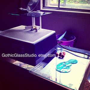 May include: A glass cutting machine with a glass platform and a glass cutting tool. The machine is on a table with a glass top. There are three small brown objects on the table. The text "GothicGlassStudio.etsy.com" is visible on the table.