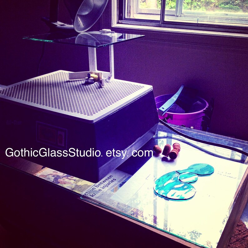 May include: A glass cutting machine with a glass platform and a glass cutting tool. The machine is on a table with a glass top. There are three small brown objects on the table. The text "GothicGlassStudio.etsy.com" is visible on the table.