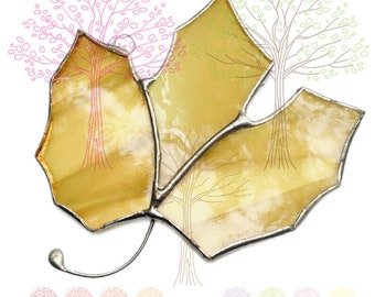May include: A stained glass leaf ornament featuring three distinct segments of translucent yellow glass held together by silver-toned metal solder lines. The leaf has a curved metal stem and a small circular loop at the top for hanging purposes.