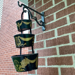 May include: A black metal planter with three tiers hangs from a hook on a brick wall. Each tier is decorated with gold accents and a different design. The planter is empty.