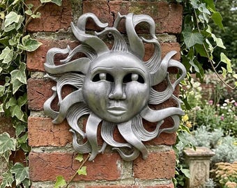 May include: A silver-colored sun face wall sculpture with flowing rays, mounted on a red brick wall. The sun has a serene expression and is surrounded by green ivy. A decorative piece for outdoor or indoor spaces.