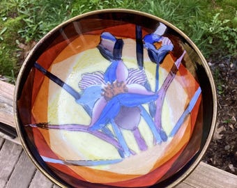 May include: A round bowl with a dark brown rim and a hand-painted interior featuring a central blue and purple flower with striped petals. The background of the bowl displays a yellow center surrounded by vibrant orange and deep red circular bands.