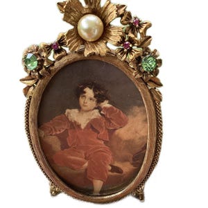 May include: Gold-toned oval picture frame with a decorative floral design. The frame is adorned with faux pearls and green and red rhinestones. The frame holds a portrait of a young boy in a red suit.