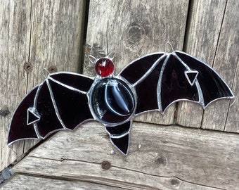May include: A stained glass bat with black and red glass. The bat has a red eye and black wings with a geometric pattern.