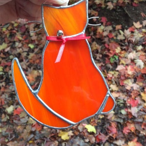May include: Orange stained glass cat with a red ribbon and a bell hanging from its neck. The cat is sitting with its tail curled around its body.