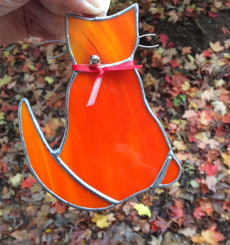 May include: Orange stained glass cat with a red ribbon and a bell hanging from its neck. The cat is sitting with its tail curled around its body.
