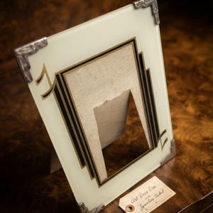 Peut inclure: Un cadre photo rectangulaire en verre avec une surface blanc cassé, des lignes de bordure géométriques noires et dorées, et des accents métalliques argentés aux coins. Une petite étiquette en papier beige porte le texte Art Deco Era et Jewelers ticket.