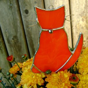 May include: Orange stained glass cat with a silver outline and a bell hanging from its neck. The cat is sitting on a bed of yellow flowers.