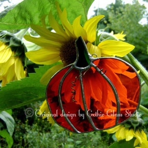Peut inclure: Un citrouille en vitrail avec une tige verte et un cadre en métal argenté est suspendue devant un tournesol. La citrouille est orange et présente un motif tourbillonnant. Le texte "Original design by Gothic Glass Studio" est visible.