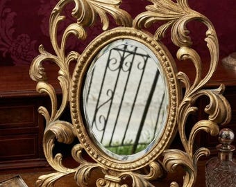 May include: Ornate gold-colored vanity mirror with an oval-shaped mirror and decorative scrollwork. The mirror is set on a wooden surface, with a small silver case and a glass perfume bottle nearby. The mirror's frame has a height of approximately 12 inches.