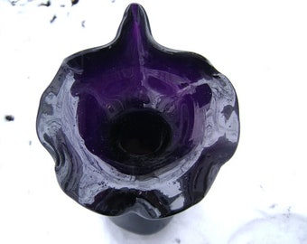May include: A dark purple glass vase with a ruffled, organic shape. The vase has a wide, fluted rim and a deep, dark interior. The glass has a glossy finish and appears to be hand-blown. The vase is a decorative item.