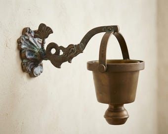 May include: A wall-mounted brass vessel holder featuring an ornate, dark-toned metal bracket with a decorative floral-shaped base plate. The swinging arm supports a cylindrical brass cup with a rounded bottom, suspended by a curved metal handle.