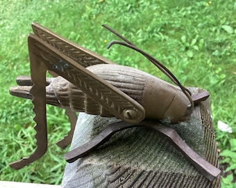 May include: A brass grasshopper figurine with a detailed design and a serrated edge on its legs. The grasshopper is standing on a metal base.