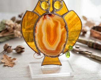 May include: A stained glass owl ornament featuring yellow glass wings and ears, two circular yellow glass eyes, and a central orange and white agate slice body. The owl is mounted on a clear acrylic base and includes a metal plaque reading Gothic Glass Studio - Handcrafted Agate Owl and the website www.GothicGlassStudio.Etsy.com.