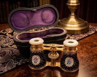 May include: Antique opera glasses with a black and gold finish, featuring floral designs and white accents. The glasses are displayed with their original case, lined with purple velvet, on a wooden surface.