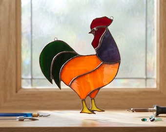 May include: A stained glass rooster suncatcher featuring a vibrant orange body, a deep purple neck, a bright red comb, and dark green tail feathers. The piece is constructed with dark metal soldering lines and includes a small metal hanging loop.