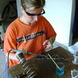 May include: A person wearing a bright orange t-shirt with black lettering is working on a stained glass project. They are using a soldering iron and a small glass cutter to create a design. The project is on a wooden table with a glass of water and a small piece of wood.