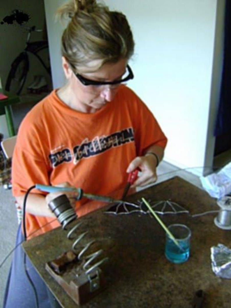 May include: A person wearing a bright orange t-shirt with black lettering is working on a stained glass project. They are using a soldering iron and a small glass cutter to create a design. The project is on a wooden table with a glass of water and a small piece of wood.
