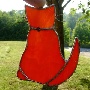May include: A stained glass cat figurine with a bright orange body and silver metal accents. The cat is sitting with its tail curled around its body.