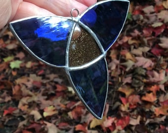 May include: A stained glass triskele ornament with a silver-colored metal frame. The three sections are a deep blue, with a central section of brown and gold. A small loop is at the top for hanging. The background is out-of-focus fall leaves.