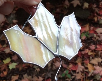 May include: A stained glass maple leaf suncatcher featuring iridescent cream and pale yellow glass panels joined by silver metal solder lines. The leaf shape includes three distinct lobes and a curved metal stem extending from the base.