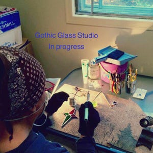 May include: A person working on a stained glass project in a studio. The person is wearing a bandana and gloves. The project is a star-shaped piece of stained glass. The studio is cluttered with tools and supplies. The text "Gothic Glass Studio In progress" is visible in the image.