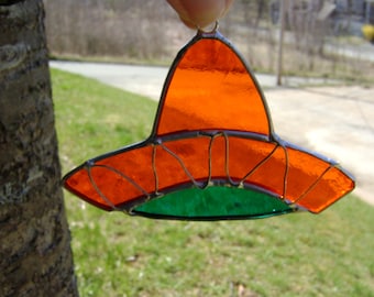 May include: A stained glass suncatcher shaped like a wide-brimmed hat, featuring vibrant orange glass panels for the crown and brim, accented with a green glass base, all held together by thin, dark metal solder lines and a small hanging loop.