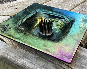 May include: A square metal dish with a raised, circular, flower frog in the center. The dish has a green, blue, and purple iridescent finish and a black butterfly design on the edge.