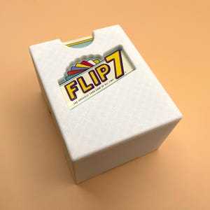 May include: A white plastic card game storage box with a textured surface. The front features a cutout window displaying the game title FLIP 7, THE GREATEST CARD GAME OF ALL TIME! in yellow, purple, and red text above a colorful fan of cards.