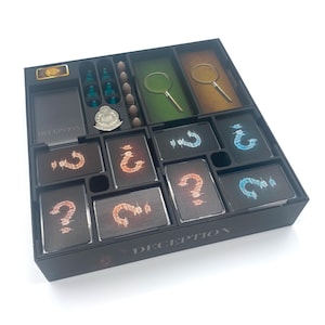 Puede incluir: Un organizador de juegos de mesa negro con compartimentos que contienen piezas de juego, cartas con signos de interrogación y fichas de lupa. La palabra "DECEPTION" está impresa en el lateral. Las piezas del juego son de varios colores.
