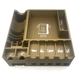 May include: A brown, multi-compartment organizer for a board game, featuring a large central area with a cat food bag graphic and the word "CATS". Smaller compartments have cat silhouettes and numbered dice icons. The organizer is designed to hold game components.