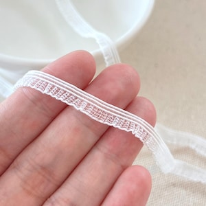 May include: A close-up shot of white lace trim with a scalloped edge. The trim is held in a hand, with a white bowl in the background. The lace appears to be made of a fine, sheer material, suitable for sewing and craft projects.