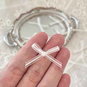 May include: A small, white bow with a lacy trim. The bow is held in a hand, with a blurred background featuring a decorative plate and white fabric. The bow is a charming accessory.